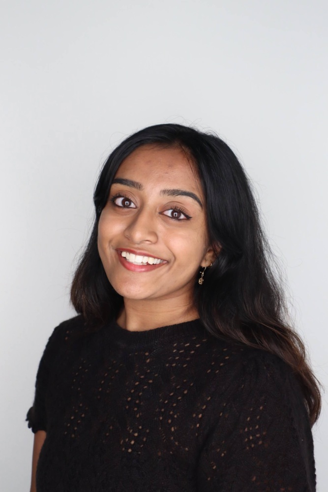A woman with long dark hair wearing a black knit top smiles at the camera against a plain light background.